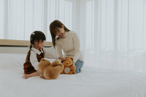 Adorable little 4-5 years old girl in headphone and mother sitting on white bed playing with bear doll toy
