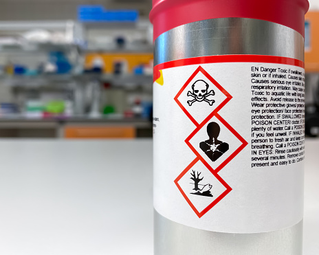 A container with a dangerous, toxic substance in a laboratory setting. Appropriate safety signs are visible on a label.