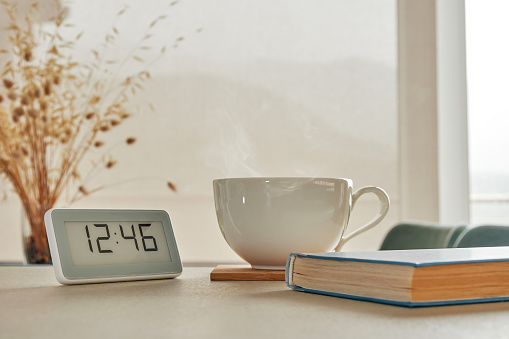 Cozy home desk with hot coffee mug, book and clocks. Wide window with beautiful mountains view