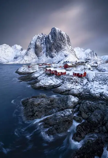 A beautiful vertical image of Lofoten Islands in Norway featuring traditional red houses or rorbuer on icy rocks during winter