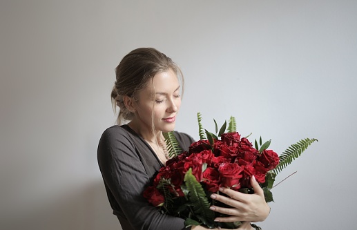 Young beautiful woman in white t-shirt with bouquet of red flowers in hands near window in bright kitchen at home