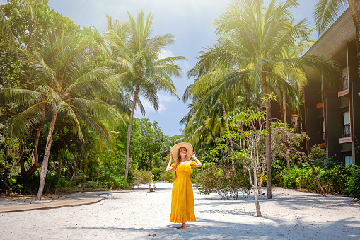 Travel woman on vacation in tropical resort. Happy young female traveler in bright yellow dress and straw hat enjoy summer holidays in luxury palm beach resort. Trip in Thailand