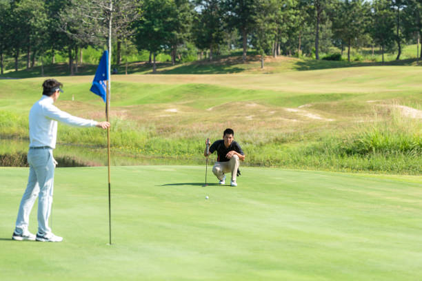 golfeur asiatique homme avec ami jouant coup de visée pour mettre la balle sur le trou avec club sur le parcours vert.  hobby en vacances et vacances en journée matinale ensoleillée sur le club de golf. concept lifestyle et sport - golf golf swing putting cheerful photos et images de collection