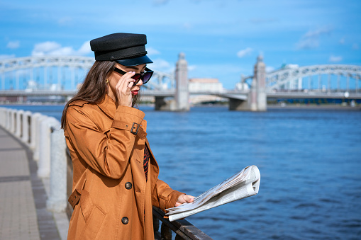 Stylish young woman of Caucasian ethnicity reads fresh newspaper in sunglasses and black cap. Beautiful brown coat standing on embankment of river on sunny day against background of sky and bridge