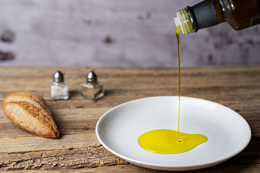 Extra virgin olive oil pouring from a bottle to a round white plate, on a rustic wooden table.