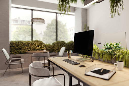 Large blank monitor with keyboard and office supplies on desk in modern office. Panoramic windows, concrete floor and green plants. Office lounge in the background. Render