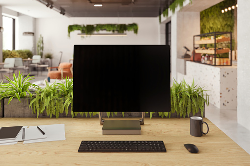 Large blank monitor with keyboard and office supplies on desk in eco-friendly office. Office cafeteria in the background. Render