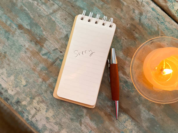istock Weathered wooden desk with a lit candle, a notepad that says “Sorry’ and pen 1439448528