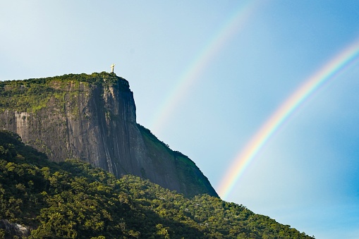 The statue of Jesus Christ on top of a cliff with the rainbow in the blue sky in Rio de Janeiro