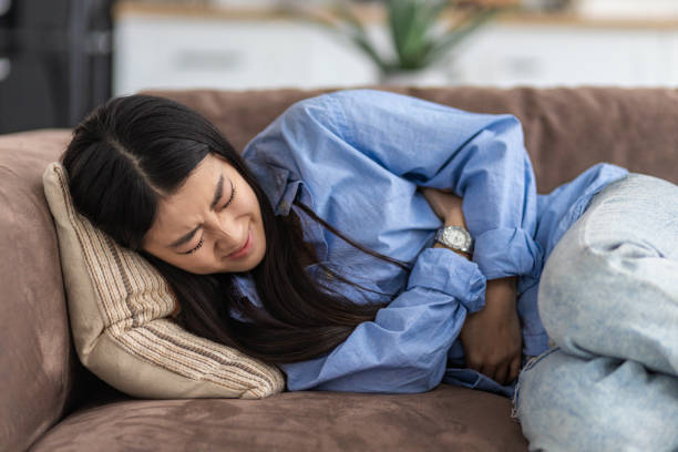 Female suffering from severe spasms holding her stomach. Young Asian woman suffering from abdominal pain lying on the couch at home stock photo