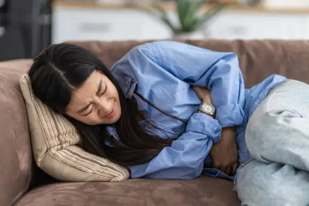 Photo of Female suffering from severe spasms holding her stomach. Young Asian woman suffering from abdominal pain lying on the couch at home