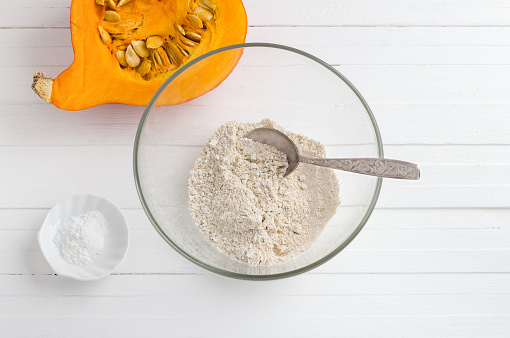 Glass bowl with oatmeal flour, half a pumpkin with seeds and baking powder on a white wooden background, top view. Cooking healthy vegan food