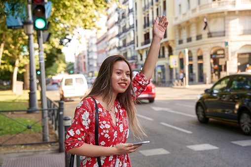 young latin woman on the street and raising arm to call to attention of a cab. She is holding a smart phone on her other hand. Concept of taxi application