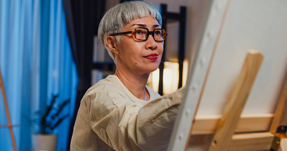 Happy Asian senior elderly woman, older people, mature lady wear glasses sit on chair looking at canvas focus art painting in workshop at night. Art therapy for seniors, Activity for elderly concept.