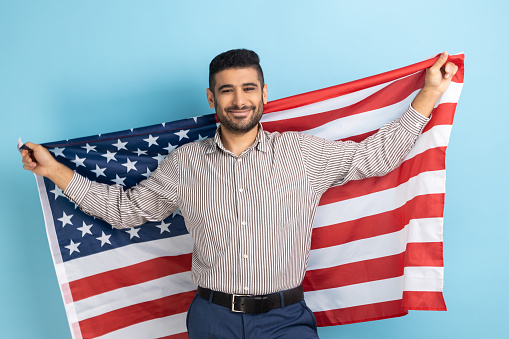 Portrait of smiling businessman holding USA flag and looking at camera with smile, celebrating national holiday, wearing striped shirt. Indoor studio shot isolated on blue background.