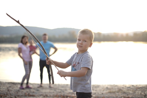 A male child playing with sticks with his family in the background behind the lake