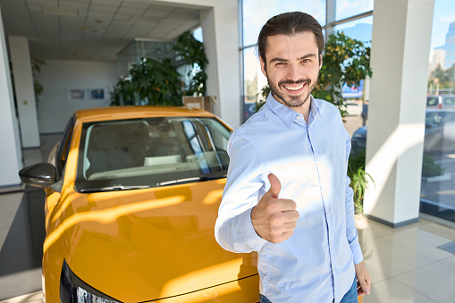 Joyous automotive dealership client standing beside new vehicle and making thumbs-up sign