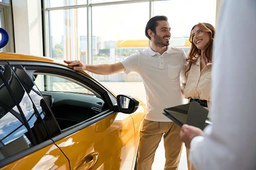 Joyous male standing beside luxury car and smiling at happy female companion in presence of salesperson