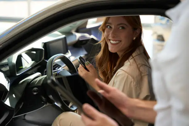 Woman with car key fob sitting at steering wheel and looking at salesclerk with tablet computer in hands