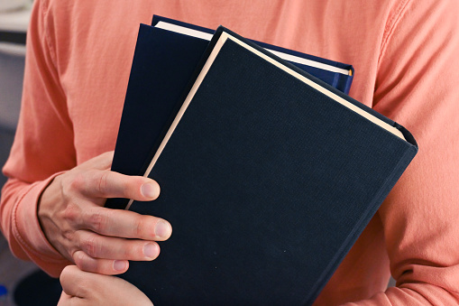 A person wearing a salmon pink sweatshirt is holding a blue and a black hardcover book in their hands.