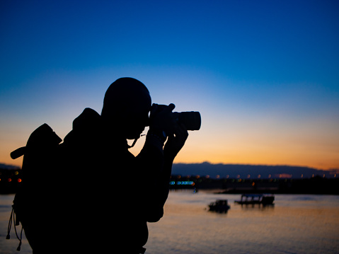 The photographer on the river bank holds the camera to his eye and takes a picture. Twilight, yellow-blue background color.