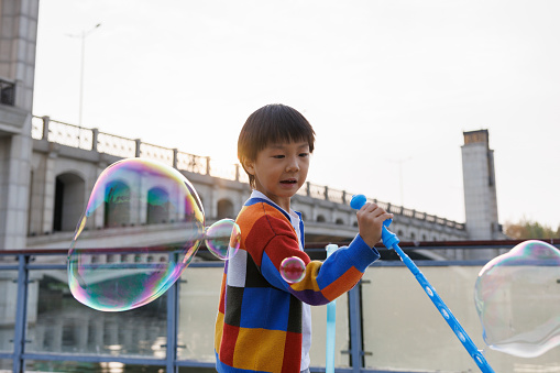 A little Asian boy, about 4 to 5 years old. He wore colorful sweaters and played with soap bubbles by the river. He was very happy. Shot by Canon R5. There is a beautiful bridge in the background.