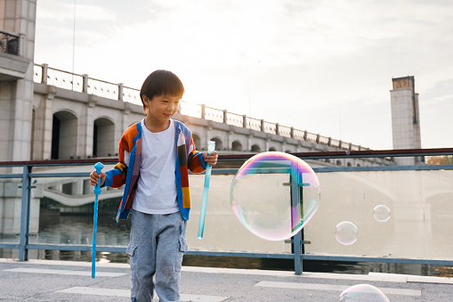 A little Asian boy, about 4 to 5 years old. He wore colorful sweaters and played with soap bubbles by the river. He was very happy. Shot by Canon R5. There is a beautiful bridge in the background.