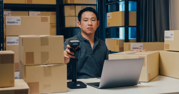 istock Asian businessman use barcode machine scan customer data in paper box and type register online information order detail in laptop for send express delivery at warehouse. 1439502450