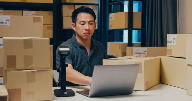 istock Asian businessman use barcode machine scan customer data in paper box and type register online information order detail in laptop for send express delivery at warehouse. 1439502452