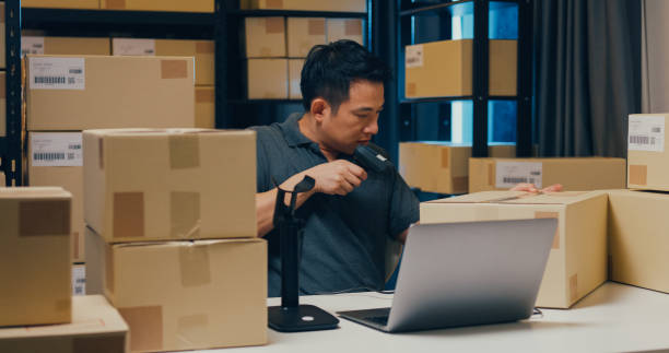 istock Asian businessman use barcode machine scan customer data in paper box and type register online information order detail in laptop for send express delivery at warehouse. 1439502458