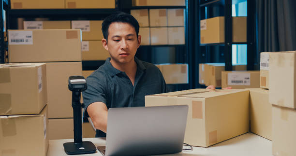 Asian businessman use barcode machine scan customer data in paper box and type register online information order detail in laptop for send express delivery at warehouse. Startup business concept. el empresario asiático utiliza la máquina de código de barras para escanear los datos del cliente en una caja de papel y escribir el registro de información en línea detalle del pedido en la computadora portátil para enviar entrega urgente en el alma