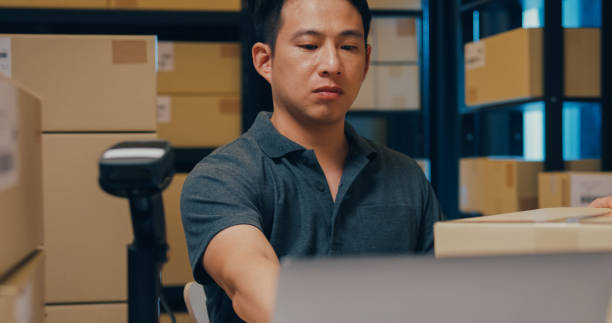 istock Close-up Asian businessman use barcode machine scan customer data in paper box and type register online information order detail in laptop for express delivery at warehouse. 1439502485