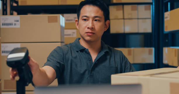 istock Close-up Asian businessman use barcode machine scan customer data in paper box and type register online information order detail in laptop for express delivery at warehouse. 1439502495