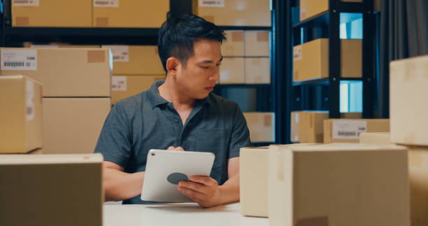 Asian businessman with barcode machine and box use digital tablet checking stock on shelf and key data in online information detail in before express delivery at warehouse. stock photo
