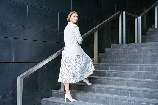 Serious mature businesswoman in elegant dress standing on stairs in front of an office building. Coach hr employee, manager, mentor motivates the employee to grow.