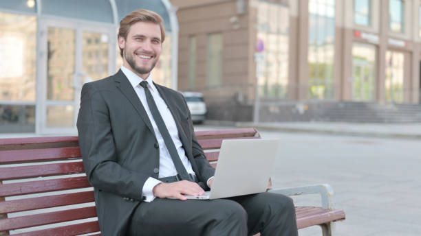 istock Businessman with Laptop Smiling at Camera while Sitting Outdoor on Bench 1439514430