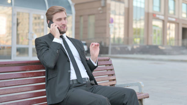 istock Businessman Talking on Phone while Sitting Outdoor on Bench 1439515552