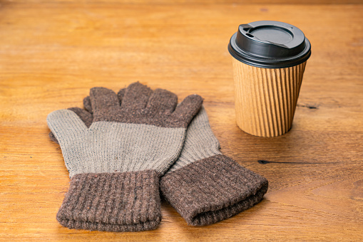 High angle view a pair of cloth gloves and a black plastic cap paper takeaway cup of hot coffee on wooden table in winter season.