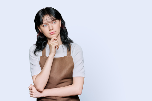 Young woman worker in apron holding hand on chin thinking thoughtfully, white background copy space place