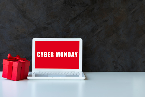 White laptop on white table  with red gift box in front of black wall . Cyber Monday text on device screen.