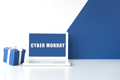 White laptop on white table with blue gift box in front of white wall with blue colored diagonal part. Cyber Monday text on device screen.