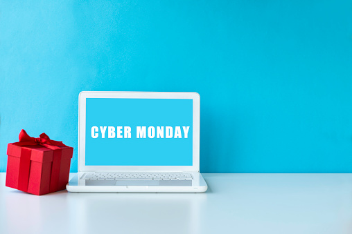 White laptop on white table  with red gift box in front of blue wall . Cyber Monday text on device screen.
