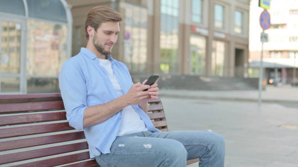 istock Man Browsing Internet on Smartphone while Sitting Outdoor on Bench 1439529409
