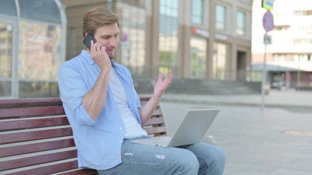 istock Angry Man Talking on Phone and using Laptop while Sitting Outdoor on Bench 1439529542