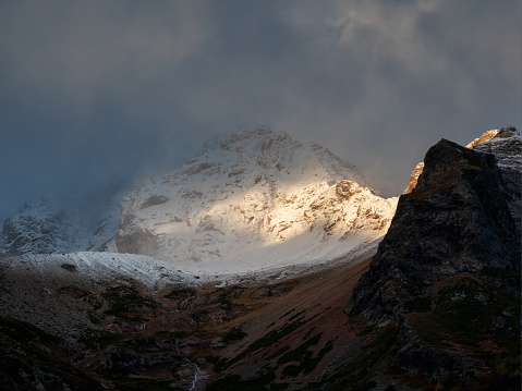 Bright ray of sunlight on a glacier. Contrasting mountains at sunrise. Dramatic mountain landscape with snowy rock in blizzard. Nature background of rocky mountain with sharp rock and cloudy sky.