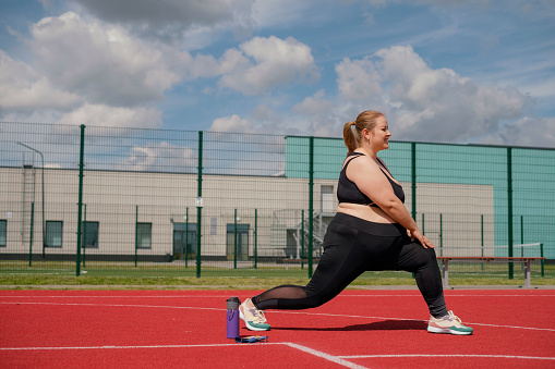 A fat obese woman does stretching and squatting on a sports field in an outdoor stadium on a hot summer morning.