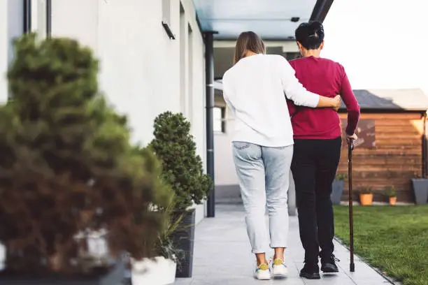 Photo of A caregiver helps an elderly woman walk in front of her house