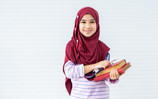 Little sweet Asian muslim girl wearing headscarf holding books for learning in class, dreaming to be doctor, smiling with happiness and looking at camera with copy space. Education, Kid Concept