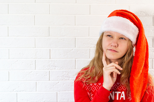 Funny photo of a teenager celebrating winter holidays, Dreamy Look, Empty Space, Think of a family party, Hand on Chin, Christmas concept in Santa hat, Red background color