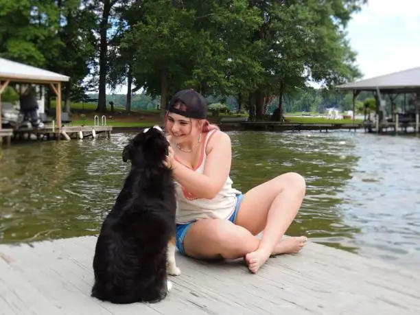 A Caucasian teenage girl in casual clothing sitting on a dock in front of a lake playing with an Australian Shepherd dog